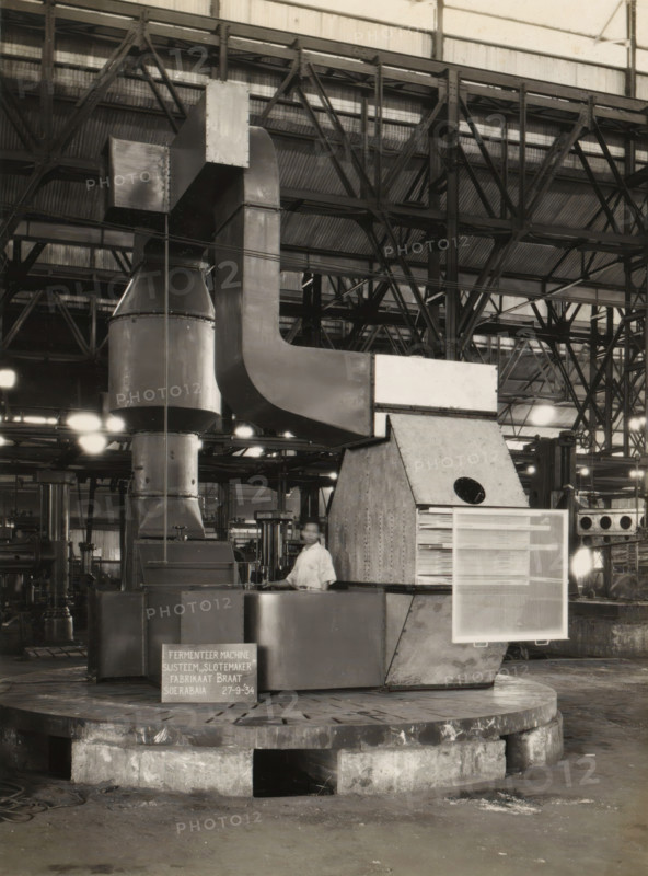 Fermentation machine, album page, photograph, A fermentation machine standing in a factory hall, September 27, 1934. Part of a photo album with photos of machines and constructions of the NV Machinefabriek Braat in Soerabaja, Soekaboemi, and Tegal from the period 1931-1937., Surabaya, 1934-09-11, photographer, Java, East Java, paper, pasteboard, gelatin silver print, developing gelatin silver print, height 240 mm x width 178 mm, height 250 mm x width 320 mm, Person, NV Machinefabriek Braat, factory-interior, machine shop, machines, parts of machines, tools and appliances, economic history, overseas history, company history, Period, 1931 - 1937, A large industrial interior with a tall metal structure composed of ducts and rectangular and curved panels rising from a rounded base, supported by a heavy circular concrete platform. The background shows a high corrugated metal roof with exposed steel trusses and beams creating a grid of diagonal and vertical lines. Bright overhead lights cast...