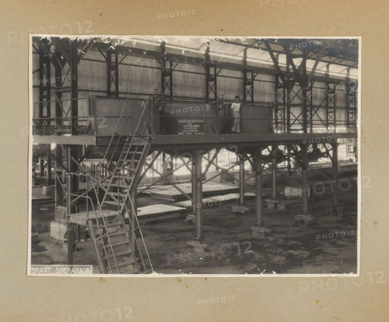 Platform with cooking pots, photograph, Album page with a photo of the interior of a factory hall containing a rack with cooking pots, ready for delivery to Guttabladbrei, October 30, 1934. Part of a photo album with photos of machines and constructions of the NV Machinefabriek Braat in Surabaya, Sukabumi, and Tegal from the period 1931-1937., Surabaya, 1934-10-30, photographer, Java, East Java, paper, pasteboard, gelatin silver print, developing gelatin silver print, height 166 mm x width 229 mm, height 250 mm x width 320 mm, Person, NV Machinefabriek Braat, factory-interior, machine shop, machines, parts of machines, tools and appliances, economic history, overseas history, company history, Period, 1931 - 1937, A sepia-toned photograph of an industrial interior showing a raised metal platform supported by a framework of girders and columns, with a set of metal stairs and handrails leading up to the platform and additional structural cross-bracing beneath; a person stands on the platf...