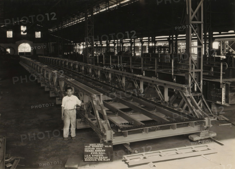 Cable bridge, album page, photograph, Album page with a photograph of a cable bridge manufactured in 1934 for the Dutch East Indies Central Procurement Service (ICA) and for installation in South Bali. Part of a photo album with photos of machines and constructions of the NV Machinefabriek Braat in Soerabaja, Soekaboemi, and Tegal from the period 1931-1937., Surabaya, 1934-12-24, photographer, Java, East Java, paper, pasteboard, gelatin silver print, developed gelatin silver print, height 174 mm x width 233 mm, height 250 mm x width 320 mm, Person, NV Machinefabriek Braat, factory-interior, machine shop, bridges by construction, economic history, overseas history, bridge construction, company history, Period, 1931 - 1937, A large indoor industrial space with high ceilings and structural beams, long metal framework assemblies arranged in parallel extending across the floor, a man standing in front of the nearest assembly wearing a light-colored shirt and trousers and resting one hand on...