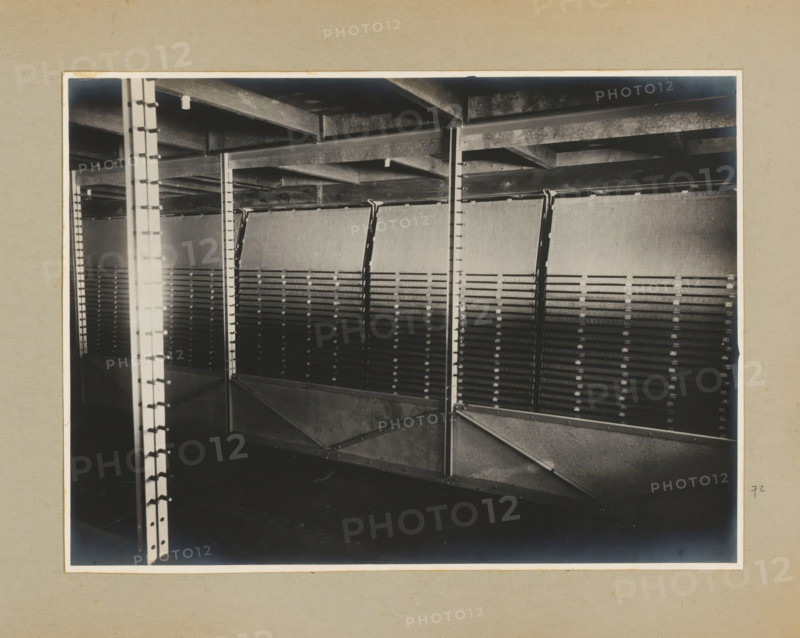 Installation, photograph, Album page with a photo of a machine or installation, inside a factory building. Part of a photo album with photos of machines and constructions of the NV Machinefabriek Braat in Surabaya, Sukabumi and Tegal from the period 1931-1937., Java, 1931 - 1937, photographer, paper, pasteboard, gelatin silver print, developed gelatin silver print, height 167 mm x width 227 mm, height 250 mm x width 320 mm, Person, NV Machinefabriek Braat, factory-interior, machine shop, economic history, overseas history, company history, Period, A black and white photograph framed by a light mat showing a metal interior structure with vertical support columns and horizontal beams, angled diagonal braces, and a series of evenly spaced slatted panels mounted between the supports, with visible rivets and bolts on the metal surfaces and a ceiling of exposed beams above, the lower area appearing dark and shadowed.