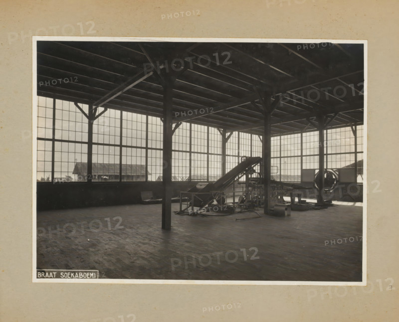 Factory hall with machine, album page, photograph, Album page with a photo of a factory building with a machine. Part of a photo album with photos of machines and constructions of the NV Machinefabriek Braat in Surabaya, Sukabumi and Tegal from the period 1931-1937., Sukabumi, 1931 - 1937, photographer, Java, West-Java, paper, pasteboard, gelatin silver print, developing gelatin silver print, height 173 mm x width 235 mm, height 250 mm x width 320 mm, Person, NV Machinefabriek Braat, factory-interior, machine shop, machines, parts of machines, tools and appliances, economic history, overseas history, company history, Period, A large interior space with high ceilings supported by dark vertical and diagonal wooden beams and a grid of horizontal ceiling joists, tall multi-pane windows lining the walls that let in light, a wooden floor with a worn parquet pattern, industrial machinery including a sloped conveyor-like apparatus and various metal components and supports clustered toward one ...