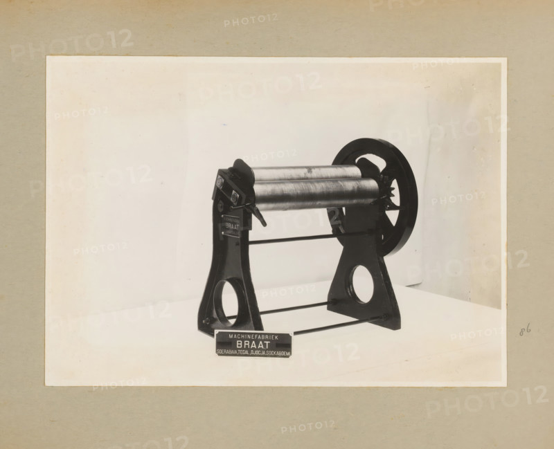 Roller press, album page, photograph, Album page with a photo of a type of roller press. Part of a photo album with photos of machines and constructions of the NV Machinefabriek Braat in Surabaya, Sukabumi, and Tegal from the period 1931-1937., Java, 1931 - 1937, photographer, paper, pasteboard, gelatin silver print, developing gelatin silver print, height 164 mm x width 230 mm, height 250 mm x width 320 mm, Person, NV Machinefabriek Braat, machines, parts of machines, tools and appliances, pressing (industrial process), economic history, overseas history, company history, Period, A photographic print of a mechanical device mounted on a plain light background, showing a pair of cylindrical metal rollers held between two dark metal A-shaped frames with circular cutouts at their bases, connected by horizontal bars; a large dark wheel with spokes is attached to one side of the rollers, and a lever-like handle is visible near the roller assembly; the image is bordered by a lighter rectangu...
