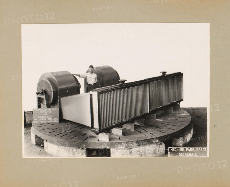 Steam engine with fan, album page, photograph, Album page with a photo of a steam engine with a fan, intended for the Reelong tea factory, June 18, 1935. Part of a photo album with photos of machines and constructions of the NV Machinefabriek Braat in Surabaya, Sukabumi, and Tegal from the period 1931-1937., Surabaya, 1935-06-18, photographer, Java, East Java, paper, pasteboard, gelatin silver print, height 162 mm x width 225 mm, height 250 mm x width 320 mm, Person, NV Machinefabriek Braat, machines, parts of machines, tools and appliances, ventilation systems, economic history, overseas history, tea culture, company history, Period, 1931 - 1937, A black and white photograph shows a large industrial machine mounted on a circular concrete base with radial slots, the machine consisting of a long rectangular radiator-like component with vertical fins and two cylindrical housings with perforated front faces, one of which has a protruding shaft. A man wearing a light-colored shirt and dark...