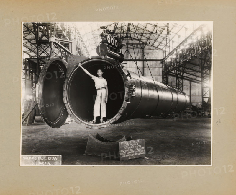 Sterilizing kettle for palm oil, album page, photograph, Album page with a photo of a sterilizing kettle for palm oil, manufactured June 22, 1935 and intended for the Sumatra Caoutchouc Maatschappij. A boy poses standing in the kettle. Part of a photo album with photos of machines and structures of the NV Machinefabriek Braat in Soerabaja, Soekaboemi, and Tegal from the period 1931-1937., Surabaya, 1935-06-22, photographer, Java, East Java, paper, pasteboard, gelatin silver print, developed gelatin silver print, height 171 mm x width 227 mm, height 250 mm x width 320 mm, Person, NV Machinefabriek Braat, factory-interior, machine shop, machines, parts of machines, tools and appliances, economic history, overseas history, palm oil cultivation, company history, Period, 1931 - 1937, A large industrial interior with a vaulted metal roof and structural steel framework contains a long cylindrical metal vessel laid horizontally across the floor. The vessel is open at one end, revealing a dark ...