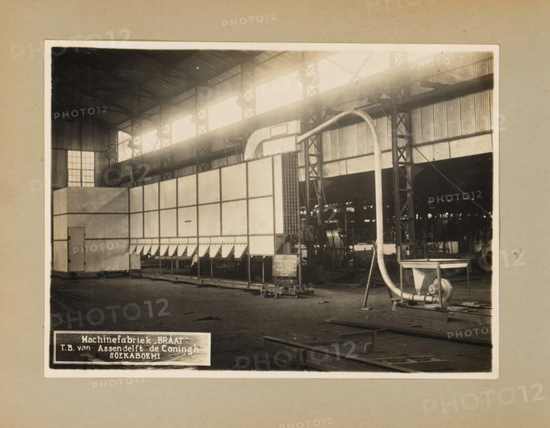 Theewan, album page, photograph, Album page with a photograph of an installation ('theewan') intended for the Kaligoea tea factory, August 6, 1935. Part of a photo album with photographs of machines and constructions of the NV Machinefabriek Braat in Surabaya, Sukabumi, and Tegal from the period 1931-1937., 1935-08-06, photographer, Java, West-Java, paper, pasteboard, gelatin silver print, developed gelatin silver print, height 172 mm x width 233 mm, height 250 mm x width 320 mm, Person, NV Machinefabriek Braat, factory-interior, machine shop, machines, parts of machines, tools and appliances, economic history, overseas history, tea culture, company history, Soekaboemi, Period, 1931 - 1937, A sepia-toned photograph of a large industrial interior showing a high roof with exposed metal trusses and tall windows along the upper wall allowing bright light to stream in, a large rectangular machine or partition with a panelled surface sits centrally and extends across much of the floor, a cur...