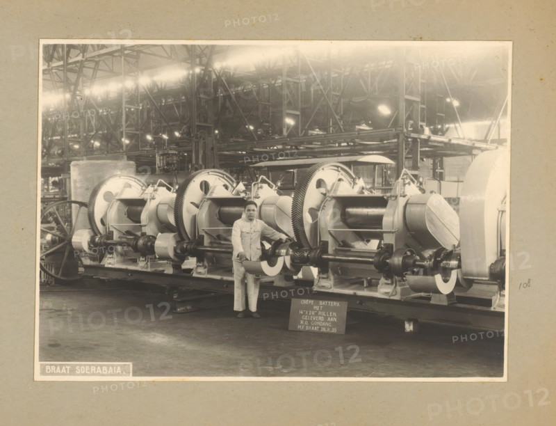 Crêpe Battery, album page, photograph, Album page with a photo of a 'crêpe battery', produced November 28, 1935 and intended for the Gondang Rubber Company. Part of a photo album with photos of machines and structures of the NV Machinefabriek Braat in Soerabaja, Soekaboemi, and Tegal from the period 1931-1937., Surabaya, 1935-11-28, photographer, Java, East Java, paper, pasteboard, gelatin silver print, developed gelatin silver print, height 160 mm x width 222 mm, height 250 mm x width 320 mm, Person, NV Machinefabriek Braat, factory-interior, machine shop, machines, parts of machines, tools and appliances, economic history, overseas history, rubber culture, company history, Period, 1931 - 1937, A sepia-toned photograph of an industrial interior showing a row of large cylindrical machines with visible gears and shafts mounted on a long base, with a man standing in front of the machinery wearing a light-colored uniform and dark shoes, metal framework and overhead trusses filling the bac...