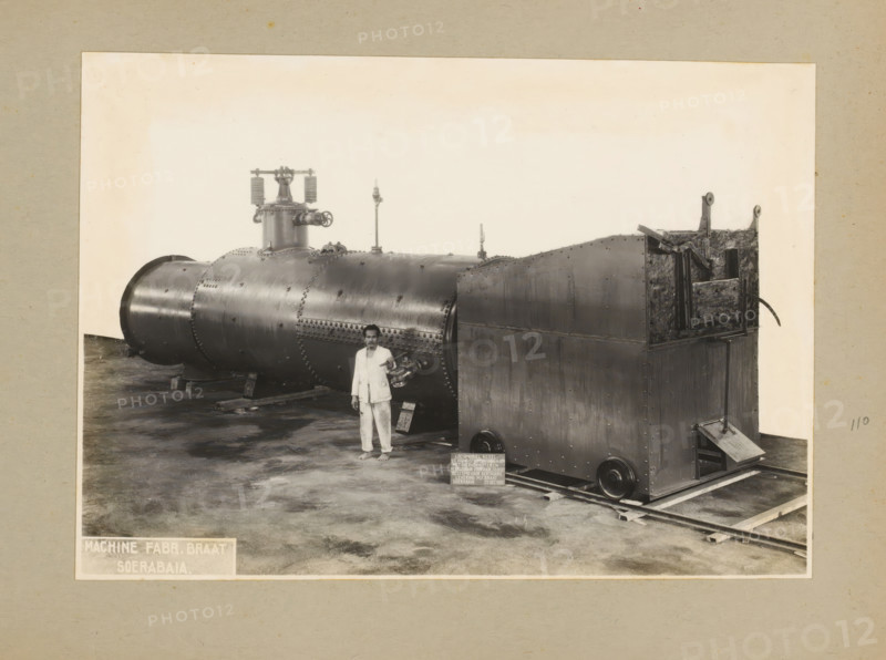 Locomobile boiler, album page, photograph, Album page with a photo of a locomobile boiler with superheater, manufactured December 23, 1935 and intended for Kertasarie. A man is posing by the boiler. Part of a photo album with photos of machines and constructions of the NV Machinefabriek Braat in Soerabaja, Soekaboemi, and Tegal from the period 1931-1937., Surabaya, 1935-12-23, photographer, Java, East Java, paper, pasteboard, gelatin silver print, developed gelatin silver print, height 160 mm x width 227 mm, height 250 mm x width 320 mm, Person, NV Machinefabriek Braat, machines, parts of machines, tools and appliances, economic history, overseas history, company history, Period, 1931 - 1937, A large riveted cylindrical metal boiler and an attached rectangular metal housing rest on wooden supports and rails inside a pale bordered frame, with a barefoot man in light-colored clothing standing in front of the cylindrical section holding a loose pipe fitting, the boiler surface showing sea...