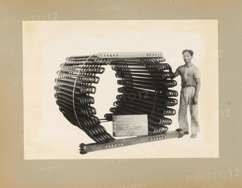 Superheater, album page, photograph, Album page with a photograph of a superheater, manufactured in 1935 and intended for (tea factory?) Kertasarie, December 23, 1935. A man poses standing next to the machine. Part of a photo album with photos of machines and constructions of the NV Machinefabriek Braat in Soerabaja, Soekaboemi, and Tegal from the period 1931-1937., Surabaya, 1935-12-23, photographer, Java, East Java, paper, pasteboard, gelatin silver print, developed gelatin silver print, height 161 mm x width 227 mm, height 250 mm x width 320 mm, Person, NV Machinefabriek Braat, machines, parts of machines, tools and appliances, economic history, overseas history, tea culture, company history, Period, 1931 - 1937, A sepia-toned photograph shows a large circular metal coil structure made of many curved rods arranged in concentric layers mounted on a horizontal base, with a man standing to the right of the structure wearing a short-sleeved shirt and loose trousers and resting one hand ...