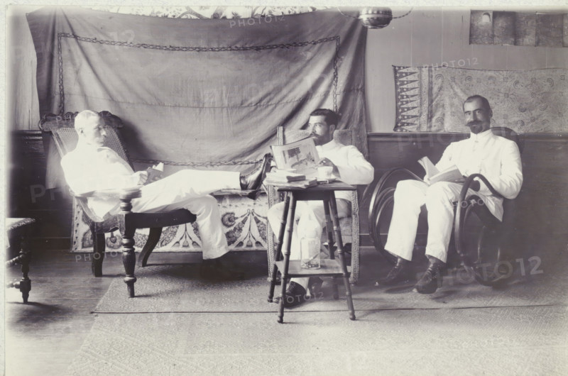 Group photo in the Gravenstraat, photograph, Group photo of Andries Boom, Dr. van Hall and Mr. van de Werk, in the house of Van Hall in the Gravenstraat in Paramaribo, October 1908. Part of the photo album of the Boom-Gonggrijp family in Suriname and Curaçao., inscription: 'Gravenstraat October 1908 P'bo', photographer: Andries Augustus Boom, Paramaribo (city), 1908-10, Andries Augustus Boom, paper, photography, height 113 mm x width 174 mm, Person, Constant Johann Jakob van Hall, overseas history, Period, 1908, Three men in light-colored clothing sit in chairs in a room with patterned fabric hung on the wall behind them, each holding a book or magazine. The man on the left reclines with his legs stretched toward the center, resting on a small table, and sits in an ornate wooden chair with a woven back. The man in the middle sits in a wicker chair, looking slightly to his left while holding an open magazine above a stack of papers on a small wooden table that has a glass bottle on its ...