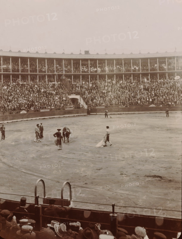At the Bull Fight, Spain, photograph, Arena for bullfights in Valencia or Barcelona., 1905 - 1922, photographer, paper, gelatin silver print, developing gelatin silver print, height 105 mm x width 85 mm, Period, A large circular arena with a dirt floor fills the image, surrounded by tall tiered wooden stands packed with people; many spectators wear hats and there are railings and arches at the front of the stands. In the arena several figures stand near a bull, including individuals in traditional clothing and capes, while a few other people are scattered around the perimeter. The foreground shows the backs of seated spectators and the top of a barrier with metal rings and posts. The overall photograph has a sepia tone and a slightly faded, aged appearance.