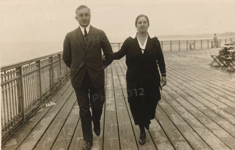 Photographs from the Estate of Isabel Wachenheimer, photograph, Eugen Wachenheimer (left) and his wife Else Wachenheimer-Moos (right) walking arm in arm on the quay of Scheveningen., date: 'Scheveningen August 1924.', Scheveningen, 1924-08, photographer, paper, gelatin silver print, developed gelatin silver print, height 90 mm x width 140 mm, Person, Eugen Wachenheimer, Else Wachenheimer-Moos, Period, A man and a woman walk side by side on a wooden boardwalk with a metal railing along the left edge; the man wears a dark three-piece suit with a white shirt and tie and has his hands behind his back, while the woman wears a dark dress or coat with a light collar and a necklace and has one arm linked through the man's arm and the other hanging by her side; the boardwalk shows parallel planks and worn patches, and in the background there are folding chairs and a distant figure near the railing with a hazy expanse beyond.