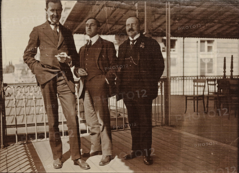 Eugen Wachenheimer as a young man with two unknown men outside under a canopy, 1906-1926, photograph, Eugen Wachenheimer as a young man with two unknown men outside under a canopy., 1906 - 1926, photographer, paper, gelatin silver print, developed gelatin silver print, height 90 mm x width 120 mm, Person, Eugen Wachenheimer, Period, Three men stand on a covered wooden balcony with a decorative metal railing. The men wear dark suits with waistcoats and ties; one man on the left leans with a hand in his pocket and the middle man holds something small between his fingers. The balcony has a slatted roof overhead and a tiled roof visible beyond the railing. In the background there are windows of a building and a couple of chairs and a small table near the railing. The photograph is sepia toned and shows fabric-like texture across the image.