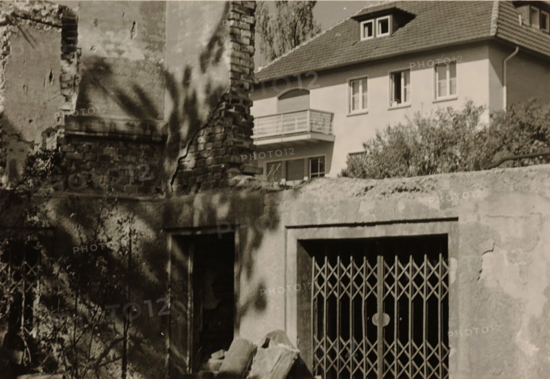 Part of a ruin with a sliding gate and a doorway in it, with a large house with a balcony in the background, with a tree and bushes around it, photograph, with a tree and bushes around it., 1945 - 1970, photographer, paper, gelatin silver print, developed gelatin silver print, height 75 mm x width 105 mm, Period, A partial view of an old brick and plaster wall with a crumbling section exposing rough bricks and a damaged window opening, shadows of foliage cast across the surfaces, a metal folding gate with a lattice pattern covering an entrance, rubble and broken masonry piled near the base, overgrown shrubs and plants along the foreground, and a neighboring house with a tiled roof, rectangular windows, and a small balcony visible behind the damaged wall.