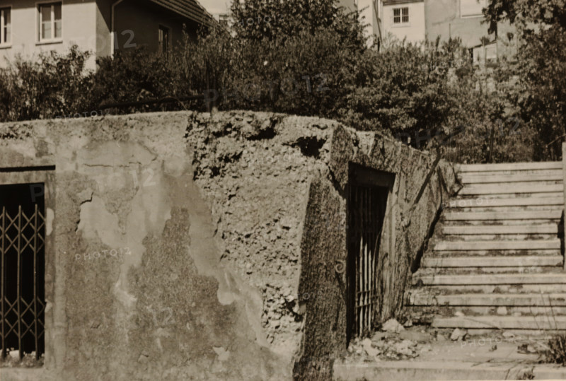 Part of a ruin with a sliding gate and a doorway in it, with a large house with a balcony in the background, with a tree and bushes around it, photograph, with a tree and bushes around it., 1945 - 1970, photographer, paper, gelatin silver print, developed gelatin silver print, height 75 mm x width 105 mm, Period, A sepia-toned scene showing a weathered concrete wall with areas of crumbling texture and peeling surface beside a set of worn stone steps leading upward; a metal barred gate set into a recessed opening is visible at the base of the wall and another barred window is set at an angle in the textured wall face; vegetation and dense shrubbery grow behind the wall, and building façades with windows and a tiled roof appear above the foliage in the background.