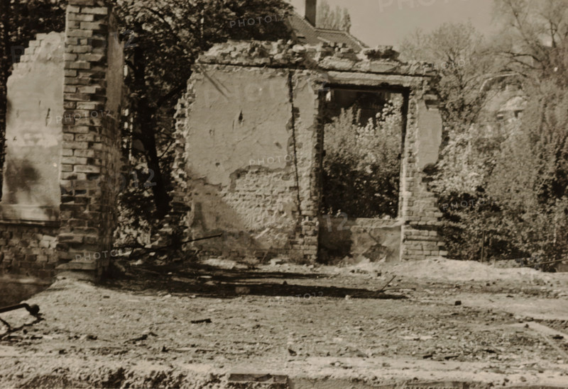 Part of a ruin, with an overgrown wall containing a doorway and bushes surrounding it, with a section of wall to the left of it, photograph, with a section of wall to the left of it., 1945 - 1970, photographer, paper, gelatin silver print, developed gelatin silver print, height 75 mm x width 105 mm, Period, A sepia-toned scene of a partially demolished brick wall with a rectangular doorway opening showing vegetation through it, rough exposed mortar and plaster on the remaining wall surfaces, scattered rubble and debris on the ground in front, a tall brick pillar at the left edge, leafy trees and bushes visible behind and around the ruined structure, and a sloped roofline of a distant building visible near the top center.