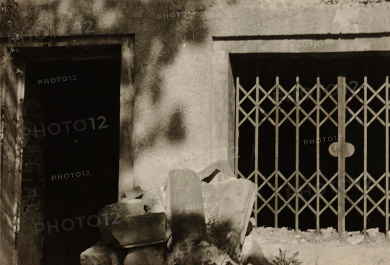 Enlargement of part of a ruin, with a sliding gate on the right and a doorway on the left, photograph, with a sliding gate on the right and a doorway on the left., 1945 - 1970, photographer, paper, gelatin silver print, developed gelatin silver print, height 75 mm x width 105 mm, Period, A cropped view of a textured wall with two rectangular openings, one dark open doorway on the left and a barred window with a diamond-pattern metal grille on the right, a pile of rough broken stones and rubble in front of the openings, a vertical column or post forming part of the grille with a round plate, and mottled light and shadow across the wall surface.