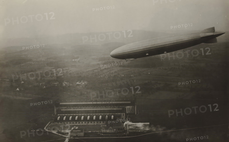 The Amerika-zeppelin ZR 3 at a hangar in Friedrichshafen, photograph, A zeppelin in the sky with a hangar on the ground and buildings in the background, inscription: 'AMERIKA-ZEPPELIN ZR III über seiner Halle in Friedrichshafen. (aufgenommen von Prof. Maerker, Konstanz)', Friedrichshafen, 1924, photographer, Baden-Württemberg, paper, gelatin silver print, developing gelatin silver print, height 85 mm x width 140 mm, Period, A large rigid airship with a streamlined body and tail fins floats above a patchwork landscape, its gondola visible beneath the hull and small windows along the side. Below the airship an industrial complex with long rectangular buildings, arched roof sections, and smaller ancillary structures sits next to open land. The surrounding area shows fields, clusters of trees, scattered houses, and gently rolling hills fading into the distance under a hazy sky.