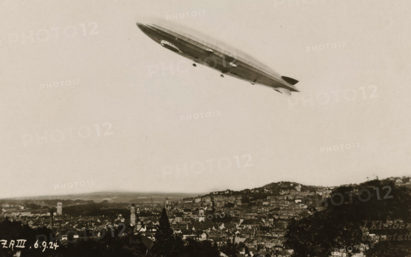 Flying Zeppelin over a city, photograph, Flying Zeppelin over a city., 1924-09-06, photographer, paper, gelatin silver print, developed gelatin silver print, height 85 mm x width 135 mm, Period, A large rigid airship floats above a sprawling cityscape, photographed from a distance; the airship has a streamlined elongated hull with a gondola underneath and small fins at the rear, shown against a pale sky. Below, the city fills the lower portion of the image with densely packed buildings, towers, and rooftops extending across rolling hills, with darker foreground trees framing the bottom edges. The overall image has a sepia tone and a slightly grainy texture.