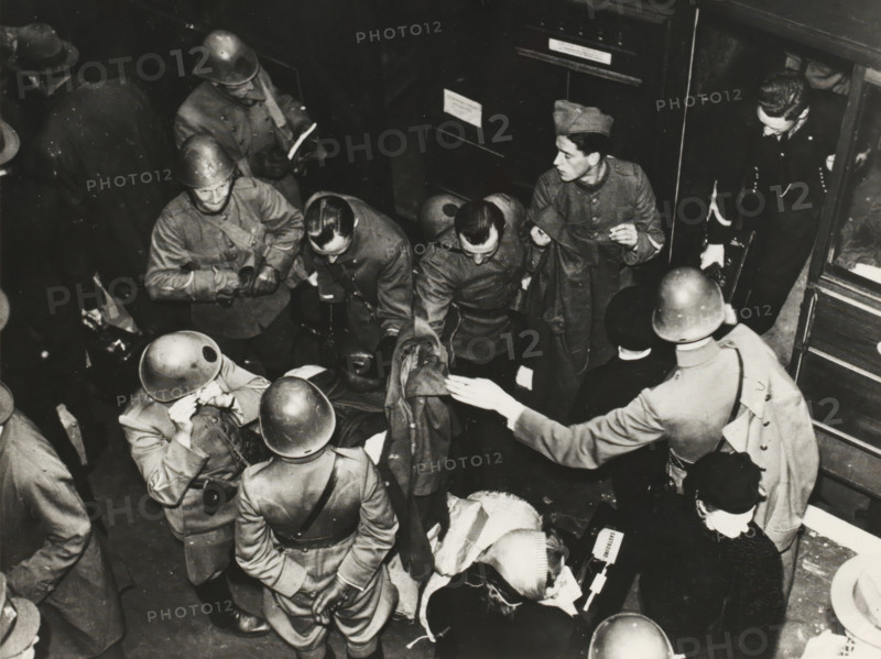 Dutch soldiers in London, photograph, Photo, taken from a certain height, with several (Dutch) soldiers stepping out of a train on a platform, with a piece of paper (carbon copy) with numbers, reverse side, with pen, explanation of those numbers: '1: Sergt. H. vd Togt, 2: Korp. J. v. Arkel, 3: HJ Brakema (Middelborst), 4: Sergt. DH Cevat, 5: Sergt. A. Domhof, 6: Private Edm. Eymael, 7: 2nd Lt. Mulders, 8: Sergt. H. Kahn, 9: 2nd Lt. Ebens, 10: Capt. L. vd Bergh, 11: Mrs. vd Bergh, 12: Mrs. Mulders., London (possibly), 1940 - 1944, photographer, London, materials by function, paper, ink, photography, writing (processes), height 14.7 cm x width 18.9 cm, Second World War, military history, England, Period, 1940 - 1945, A crowded black-and-white scene showing several uniformed men wearing helmets and coats gathered closely together, some facing inward and some turned toward the edges of the frame, with a few men bending over or reaching toward items on a surface; one man at right wears a di...