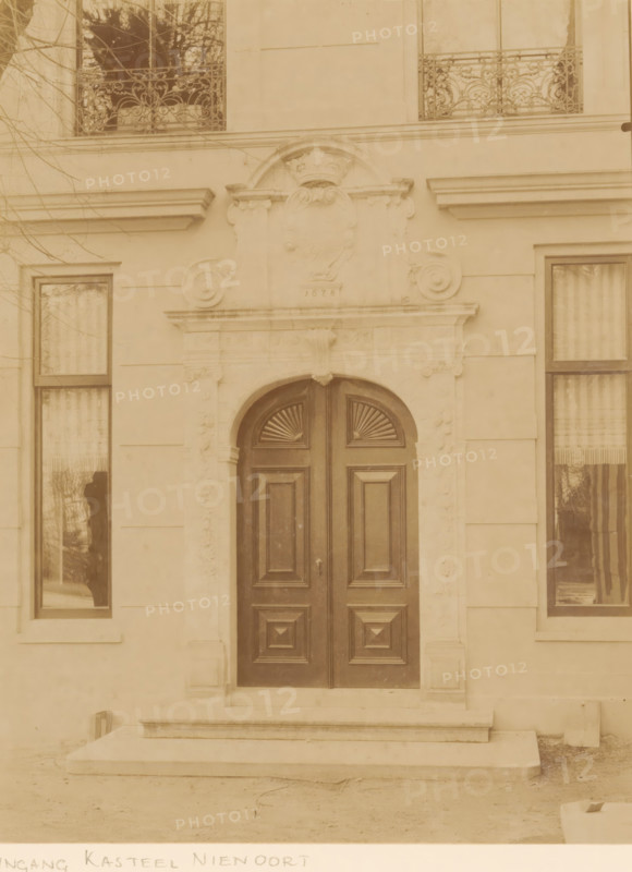 Portal of Nienoord Manor in Leek, photograph, annotation: 'Entrance of the castle (modern), Nienoord, in Leek province (Gr.), the small gate from 1678 has apparently been restored, April 1897', annotation: 'Entrance Nienoord Castle, Leek', Leek, 1897, photographer, Attributed to, paper, pasteboard, albumen print, height 228 mm x width 174 mm, gate, entrance, A double wooden door with carved rectangular panels and fan-shaped carvings in the upper sections sits within an arched stone doorway that has ornate carved details and a decorative crest above, flanked by tall windows with curtains and reflective glass, stone steps leading up to the entrance, smooth stone facade with horizontal joint lines, and parts of bare tree branches visible against the building.