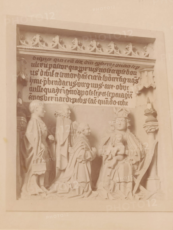 Bas-relief in the St. Hippolytus Church in Middelstum, photograph, annotation: 'Bas-relief in the Dutch Reformed Church in Middelstum', annotation: 'Middelstum', St. Hippolytus Church, 1890 - 1910, photographer, Middelstum, Attributed to, paper, pasteboard, albumen print, height 223 mm x width 171 mm, sculpture (relief, sculpture), A carved stone relief framed by a rectangular border shows a group of robed figures arranged in a scene, with one seated figure wearing a crown and holding a small child on their lap, flanked by standing and kneeling figures in long draped garments; above them is a horizontal panel with ornate floral trim and rows of stylized script carved into the stone, and decorative architectural elements appear at the top corners and sides.