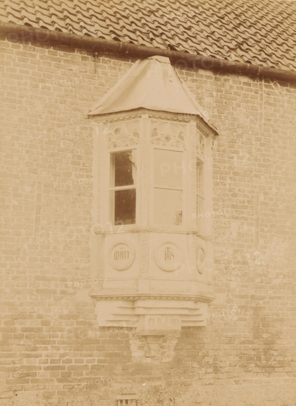 Bay window on the north facade of the Ter Apel Monastery, photograph, annotation: 'Ter Apel', annotation: 'Ter Apel, Bay window on the north facade of the monastery, Sept 1893', Ter Apel, 1893, photographer, Attributed to, paper, pasteboard, albumen print, height 232 mm x width 170 mm, bay-window, oriel, A sepia-toned photograph of a brick wall with a projecting bay window feature topped by a small pitched roof, the bay window has decorative carved panels above the glass panes and circular carved medallions below, a gutter and tiled roof run along the top of the image, and the brickwork shows varied tones and weathering.