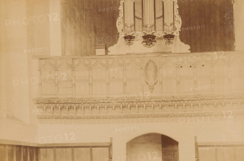 Choir screen in the Monastery Church Ter Apel, photograph, annotation: 'Ter Apel', annotation: 'Ter Apel, Jubilee in the church, total view, Sept 1893', Ter Apel, 1893, photographer, Attributed to, paper, pasteboard, albumen print, height 158 mm x width 235 mm, interior of church, rood-screen, choir-screen, iconostasis, A sepia-toned photograph showing an interior wall with decorative wooden paneling and ornate carved stonework; at the top center there is a section of an organ with visible vertical pipes and carved scrollwork beneath them, flanked by dark wooden panels. Below the organ a high balcony-like wooden railing features repeated carved panels with pointed arch motifs and a central carved niche with a sunburst or shell-shaped relief. Beneath the railing a horizontal band of intricate stone tracery and rounded quatrefoil-like patterns runs across the wall. Lower on the wall an arched opening leads into a recessed space, and wooden cupboard doors or panels line the lower portions...