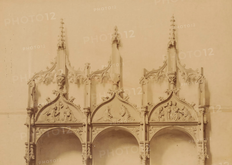 Details of the three stone seats in the Kloosterkerk Ter Apel, photograph, annotation: 'Ter Apel, Upper part of the 3 stone seats against the south wall of the choir of the church, Sept 1893', annotation: 'Ter Apel', Ter Apel, 1893, photographer, Attributed to, paper, pasteboard, albumen print, albumendruk, height 164 mm x width 231 mm, interior of church, sculpture (relief, sculpture), chair (stone), A carved stone architectural panel with three pointed ornamental gables sitting atop a series of arches, each gable containing shallow relief scenes of human figures in various poses; slender pinnacles rise above each gable with decorative crockets, tracery and small sculpted floral motifs along the vertical supports, and columns with capitals separating the arches below; the surface has a uniform warm sepia tone and shows slight variations in shading and some small speckled marks.