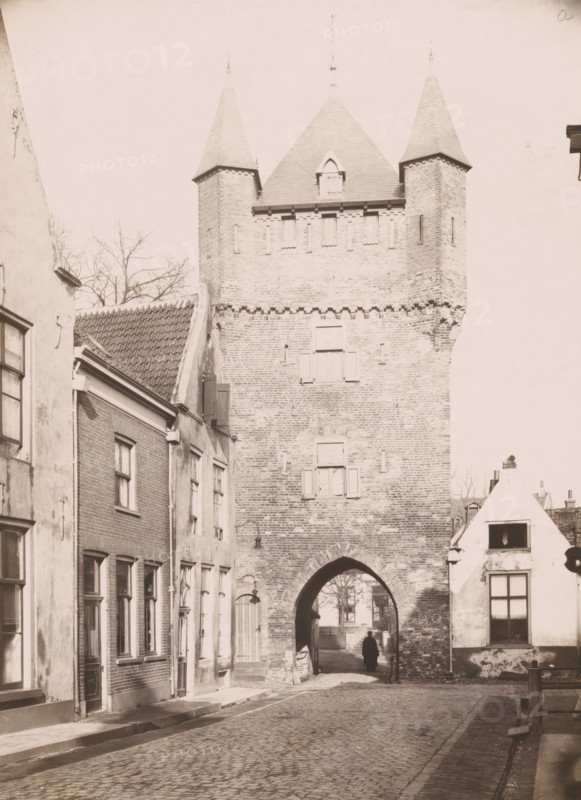 View of the Dijkpoort in Hattem, photograph, annotation: 'a, Hattem', annotation: 'Hattum, Dijkspoort in restored condition Feb. 1910', Hattem, 1910, photographer, Attributed to, paper, pasteboard, daylight collodion silver print, height 218 mm x width 159 mm, city-gate (city(-scape) with figures, staffage), Dijkpoort, A tall brick gatehouse with two pointed turrets and a steep central roof rises above a cobbled street, showing small shuttered windows and an arched passage at its base; flanking the gatehouse are rows of narrow brick and plaster buildings with tiled roofs, rectangular windows, doors, and wall-mounted lamps; a solitary figure stands in the shadowed archway, and leafless tree branches are visible behind the buildings.