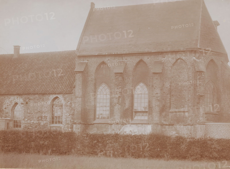 View of the choir of the Reformed Church in Andelst, photograph, annotation: 'Church of the Dutch Reformed congregation in Andelst, Gelderland, Nov 1910, Municipality of Valburg', Andelst, 1910, photographer, Attributed to, paper, pasteboard, daylight collodion silver print, height 158 mm x width 217 mm, parts of church exterior and annexes: choir, A sepia-toned photograph of a large brick building with steep tiled roofs and arched windows, showing textured brickwork and stone patches on the lower wall, a row of tall pointed-arch windows with multi-pane glass, a smaller tiled roof section with a chimney, and a hedge running along the base of the building with a grassy area in the foreground.