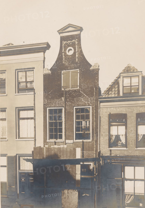 Façade of Langestraat 102 in Amersfoort, photograph, annotation: 'Amersfoort (U.), Langstraat No 102, Oct 1902, Aug 1902', Amersfoort, 1902, photographer, Attributed to, paper, pasteboard, gelatin silver print, height 217 mm x width 148 mm, urban housing, façade (of house or building), A narrow brick building with a curved stepped gable and a triangular pediment at the top stands between adjacent buildings, featuring a small circular opening near the top and decorative stone scrolls on either side of the gable, tall multi-pane windows on the lower floors with shutters on the upper window, and visible brick texture and weathering; neighboring facades include a lighter smooth-stucco building with rectangular windows and blinds on the left and a building with tiled roof dormer and windows with curtains on the right, while wooden boards and a dark awning or canopy cover part of the lower frontage.