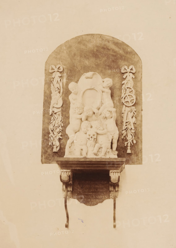 Tomb monument of Jacob van Campen in the St. George's Church in Amersfoort, photograph, annotation: 'Amersfoort', annotation: 'Amersfoort, S. Joriskerk, ... in memory of Jacob van Kampen obiit 1657', St. George's Church (Amersfoort), 1891, photographer, Amersfoort, Attributed to, paper, pasteboard, albumen print, height 222 mm x width 164 mm, Person, Jacob van Campen, other memorial structures, A carved stone relief mounted on a wall with an arched dark backdrop shows a group of cherubic figures clustered around a central oval cartouche, with some figures sitting and others standing; one figure holds a small skull near the center. Flanking the relief on both sides are vertical decorative garlands tied with bows at the top, each garland incorporating ribbonlike elements and ornamental shapes. The relief rests on a projecting stone shelf supported by carved brackets, with two curved metal supports hanging down from the shelf. The overall image has a sepia tone and shows the wall surround...
