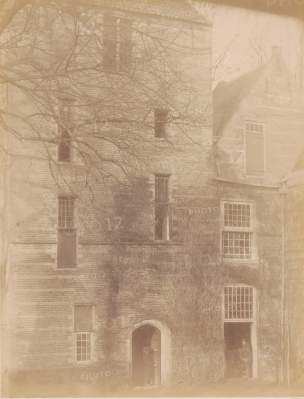 Part of IJsselstein Castle, photograph, annotation: 'IJsselstein', annotation: 'Castle in IJsselstein, demolished 1888', IJsselstein, c. 1888, photographer, Attributed to, paper, pasteboard, albumen print, height 233 mm x width 178 mm, ruin of a dwelling, house, castle, landscape with ruins (city(-scape) with figures, staffage), IJsselstein Castle, A faded photograph of a brick building facade with multiple windows and doorways, bare tree branches extending across the upper portion, a rounded arch doorway near the lower center with a person standing inside the shadowed opening, a taller rectangular opening beside it with another person partly visible in darkness, several rectangular windows with grid-like panes and some shuttered openings, a gabled roof section on the right, and climbing vines or vegetation covering parts of the lower and middle walls.