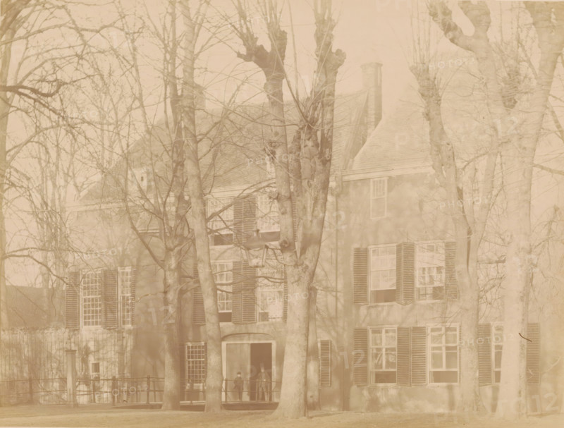 View of IJsselstein Castle, photograph, annotation: 'Castle in IJsselstein, demolished 1888', annotation: 'IJsselstein', IJsselstein, c. 1888, photographer, Attributed to, paper, pasteboard, albumen print, height 174 mm x width 230 mm, ruin of a dwelling, house, castle, landscape with ruins (city(-scape) with figures, staffage), IJsselstein Castle, A faded photograph of a large building partially obscured by many leafless trees, showing a facade with shuttered windows, a pitched roof with a chimney, and a doorway with a small porch; two standing figures are visible on the porch beneath the trees; a streetlamp is positioned near the center, and a low fence runs along the front of the building.