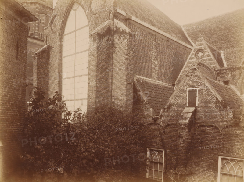 South side of the Old St. Nicholas Church in IJsselstein, photograph, annotation: 'Church in IJsselstein, South side, tower, transept, consistory, Nov. 1890', annotation: 'IJsselstein', IJsselstein, 1890, photographer, Attributed to, paper, pasteboard, albumen print, height 177 mm x width 233 mm, church (exterior), Oude Sint-Nicolaaskerk, A sepia-toned view of an old brick building complex showing a large arched window with a grid of panes set in a tall wall, adjacent buttressed brickwork with weathered and mossy patches, steep tiled roofs, a small gabled section with a dark shuttered window framed by lighter trim, decorative brick patterns on a triangular gable, arched openings beneath a row of roof tiles, two arched glazed windows lower on the wall, and dense foliage and branches growing in front of the lower portion of the walls.