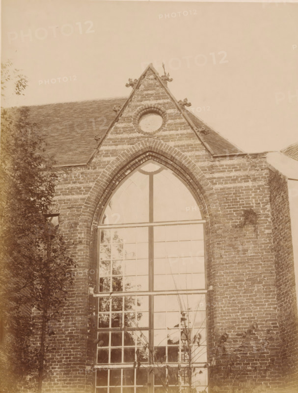 Part of the choir of the Old St Nicholas Church in IJsselstein, photograph, annotation: 'Church in IJsselstein, Unplastered ... of the choir, Nov. 1890', annotation: 'IJsselstein', IJsselstein, 1890, photographer, Attributed to, paper, pasteboard, albumen print, albumendruk, height 230 mm x width 176 mm, parts of church exterior and annexes: choir, Oude Sint-Nicolaaskerk, A section of a brick building with a steep gabled roof and decorative stone accents under the gable peak, a circular masonry feature near the top of the gable, and a tall arched window with multiple panes occupying the central facade; portions of climbing plants and tree branches frame the left side and base of the wall, and the brickwork shows weathering and mortar lines.