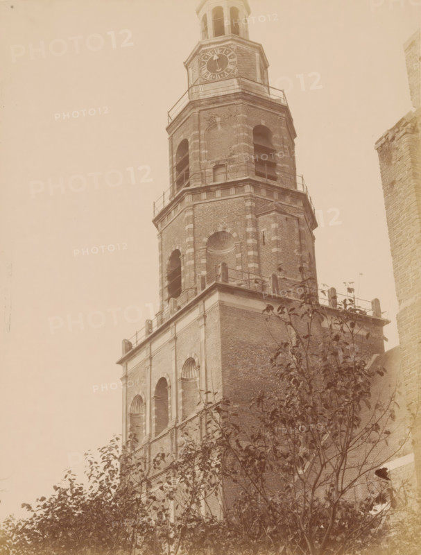 Tower of the Old St. Nicholas Church in IJsselstein, photograph, annotation: 'Tower in IJsselstein, south-east side, (upper part), Nov. 1890', annotation: 'IJsselstein', IJsselstein, 1890, photographer, Attributed to, paper, pasteboard, albumen print, height 231 mm x width 175 mm, parts of church exterior and annexes: tower, Oude Sint-Nicolaaskerk, A tall brick tower with multiple arched openings and decorative brickwork rises into the sky, showing a section with a clock face near the top and an open belfry above it, metal railings encircling several tiers, and a portion of a neighboring brick wall visible at the right edge; in the foreground leafy tree branches partially obscure the lower part of the tower.
