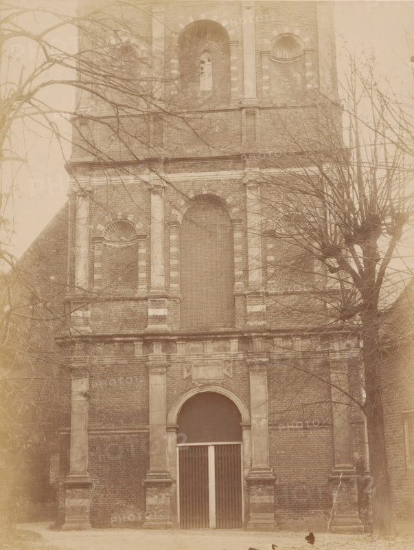 Tower of the Old St. Nicholas Church in IJsselstein, photograph, annotation: 'IJsselstein', annotation: 'Tower in IJsselstein, Westfront (lower part), Nov. 1890', IJsselstein, 1890, photographer, Attributed to, paper, pasteboard, albumen print, height 232 mm x width 176 mm, parts of church exterior and annexes: tower, Oude Sint-Nicolaaskerk, A tall brick building facade with stone columns and decorative arches, leafless tree branches partially obscuring the view, a large arched doorway with vertical slatted doors, recessed niches with shell-shaped reliefs, rectangular stone detailing around windows and along the facade, and bare ground in front of the entrance.