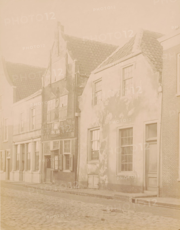 Houses in the Benschopperstraat in IJsselstein, photograph, annotation: 'IJsselstein', annotation: 'House on Benschopperstraat in IJsselstein demolished 1889', IJsselstein, c. 1889, photographer, Attributed to, paper, pasteboard, albumen print, height 231 mm x width 179 mm, urban housing, street, A row of old buildings lines a cobbled street, showing varied facades and rooflines with tiled roofing; one building has a stepped gable and decorative brickwork, another has a plastered exterior with patches of peeling paint and several multi-pane windows, and a narrow doorway set into its wall; wooden window shutters appear open on one facade, while stone and brick details are visible around entrances and lower walls; the street foreground shows uneven cobblestones and a raised pavement edge.