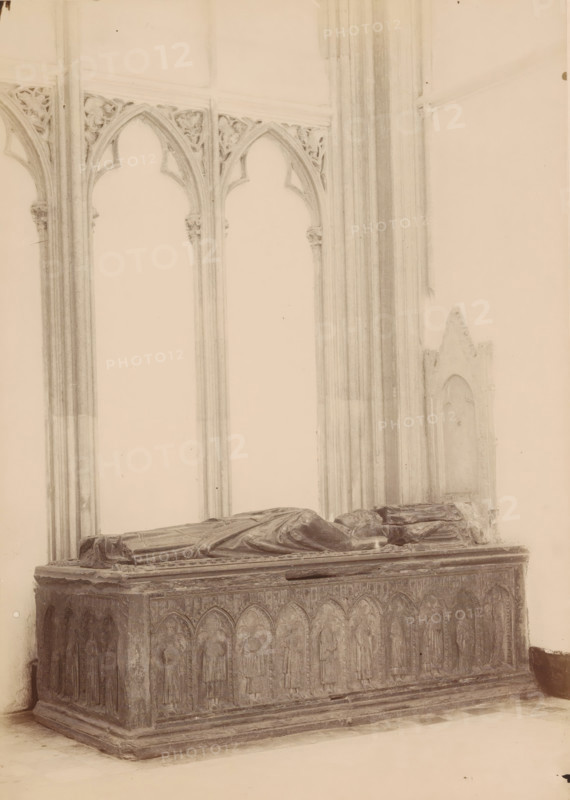 Tomb monument of Gwijde van Avesnes in the Cathedral of Utrecht, photograph, annotation: 'Utrecht', annotation: 'Cathedral in Utrecht, tomb of Bishop Guy van Avesnes, Sept 1891', Cathedral of Utrecht, 1891, photographer, Utrecht, Attributed to, paper, pasteboard, albumen print, height 240 mm x width 171 mm, Person, Gwijde van Avesnes, interior of church, grave-building, monumental tomb, A stone tomb with a rectangular base and carved arched panels along its sides occupies the foreground, topped by two recumbent effigies draped in robes. Behind the tomb rise tall, slender columns and pointed arch openings with ornate tracery in the spandrels, and a section of a wall with a recessed arched niche is visible to the right. The scene is rendered in muted monochrome tones with soft lighting and subtle shadows.