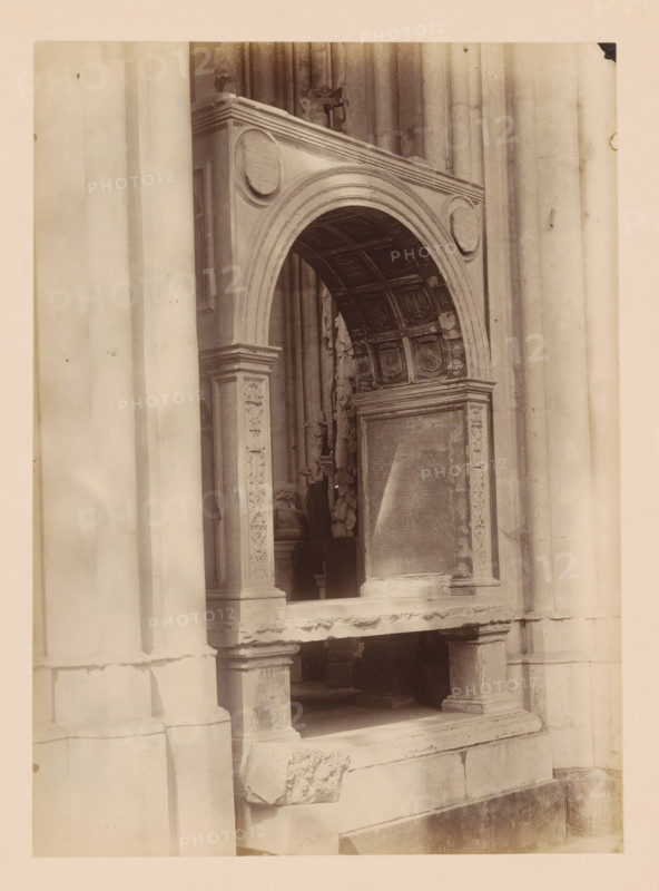 Grave monument in the Dom of Utrecht, photograph, annotation: 'Utrecht', annotation: 'Cathedral in Utrecht, grave monument, Sept 1891', Dom of Utrecht, 1891, photographer, Utrecht, Attributed to, paper, pasteboard, albumen print, height 239 mm x width 173 mm, interior of church, grave-building, monumental tomb, A sepia-toned photograph of a stone architectural feature showing a carved archway with decorative panels and reliefs; the arch has an ornate coffered underside with square recessed panels and shield-like motifs, and circular medallions set into the outer corners; fluted columns and pilasters with carved floral and geometric ornament flank the opening, and a recessed rectangular panel with worn surface sits beneath the arch; the stone shows weathering and surface irregularities, and a ledge and base with visible joints and chipped edges occupy the lower portion of the image.