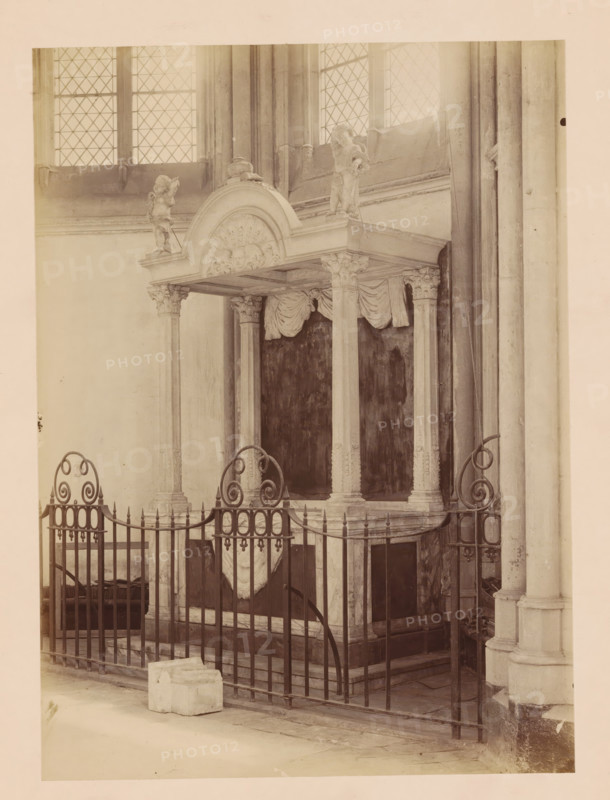 Grave monument in the Dom of Utrecht, photograph, annotation: 'Domkerk te Utrecht, Monument in der kooromgang, Sept 1891', annotation: 'Utrecht', Dom of Utrecht, 1891, photographer, Utrecht, Attributed to, paper, pasteboard, albumen print, albumendruk, height 325 mm x width 171 mm, interior of church, grave-building, monumental tomb, A stone canopy supported by carved columns stands behind an ornate wrought iron fence; the canopy features a rounded pediment with sculpted decorative relief and sculpted figures at its corners, draped fabric hanging beneath its roof, and a dark recessed background beneath the structure; tall leaded windows with diamond panes are visible above, and a stone column with vertical fluting appears at the right edge; a small block of stone rests on the floor in front of the fence.