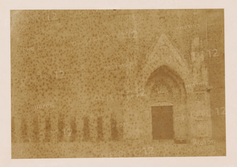 West wall of the cloister at the Dom of Utrecht, photograph, annotation: 'Utrecht', annotation: 'West wall of the cloister in Utrecht', Dom of Utrecht, 1890 - 1910, photographer, Utrecht, Attributed to, paper, pasteboard, albumen print, height 161 mm x width 232 mm, cloisters, monastery, A faded sepia photograph showing a textured brick wall with darker spotting across its surface and a pointed stone archway set into the wall on the right side, the archway featuring carved decorative stonework and a recessed doorway with a dark wooden door, flanking stone columns and sculpted relief above the arch, with part of a pale border visible around the image.