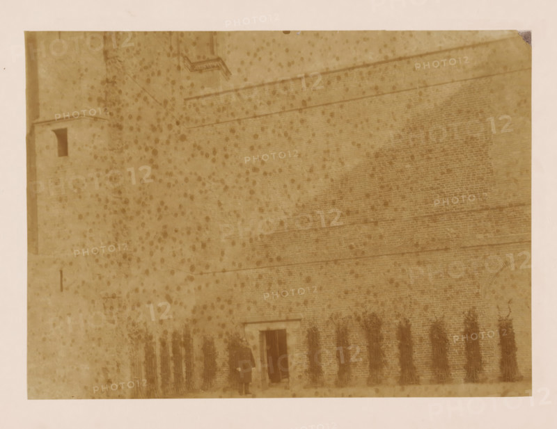 West wall of the cloister at the Dom of Utrecht, photograph, annotation: 'West wall of the cloister in Utrecht, Dec 1900', annotation: 'Utrecht', Dom of Utrecht, 1900, photographer, Utrecht, Attributed to, paper, pasteboard, albumen print, height 160 mm x width 220 mm, cloisters, monastery, A sepia-toned photograph of a large brick wall with faint horizontal lines and a small doorway near the bottom center, flanked by a row of tall, bundled vertical objects against the wall, with a person standing near the doorway wearing a long dark coat and a hat; the surface of the image shows numerous dark speckled spots and a lighter border around the edges.