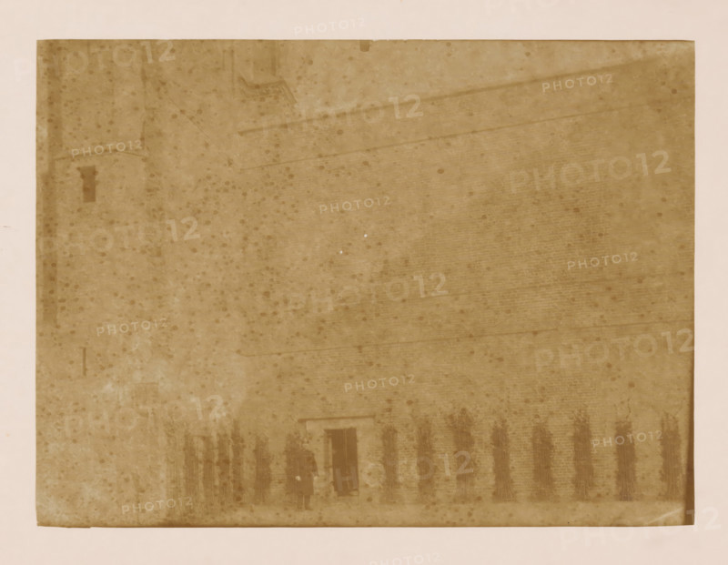 West wall of the cloister at the Dom of Utrecht, photograph, annotation: 'West wall of the cloister in Utrecht (Utrecht), Dec 1900', annotation: 'Utrecht', Dom of Utrecht, 1900, photographer, Utrecht, Attributed to, paper, pasteboard, albumen print, height 170 mm x width 230 mm, cloisters, monastery, A faded sepia-toned rectangular photograph with a wide lighter border shows a large brick wall and a sloped roof occupying most of the frame, the surface marked by scattered round spots and subtle streaks; at the lower center a dark doorway is visible with two faint silhouetted figures standing nearby, and a row of dark vertical bundles or stakes leans against the wall to the right of the doorway.