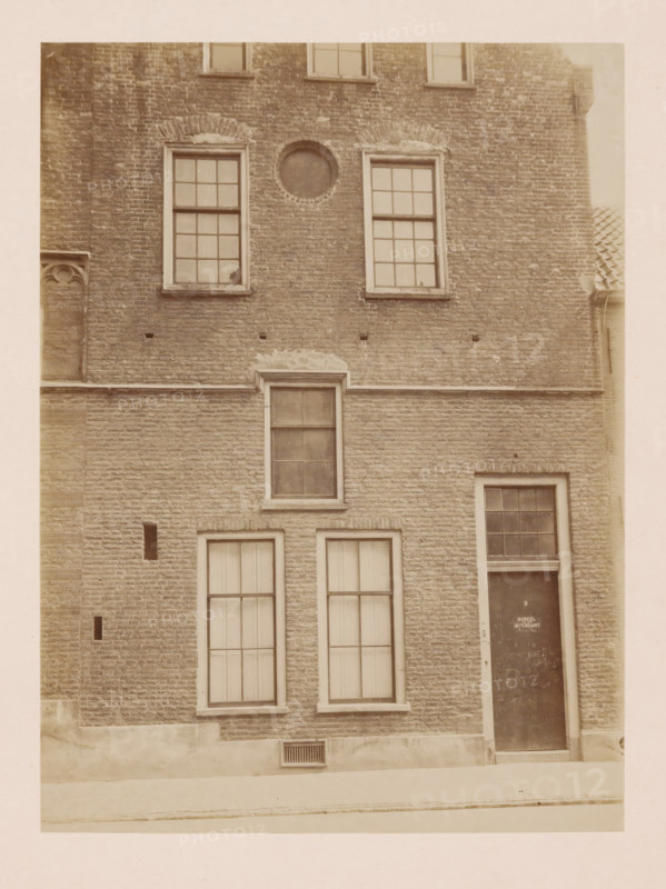 Part of the former Agnieten Convent in Utrecht, photograph, annotation: 'Utrecht', annotation: 'Utrecht, Former St. Agnieten Convent now military building, part bordering a, d church, June 1902', Utrecht, 1902, photographer, Attributed to, paper, pasteboard, albumen print, height 230 mm x width 170 mm, cloisters, monastery, A brick building facade with multiple rectangular windows framed in light trim, a circular recessed opening above two upper windows, a doorway with a darker door set into the brick, narrow vertical vents in the lower brick, a horizontal stone band separating upper and lower sections, a small rectangular grille near the base, and a tiled roof edge visible at the right.