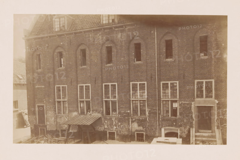 View of the former Agnieten Convent in Utrecht, photograph, annotation: 'Utrecht', annotation: 'Utrecht (U.), Former St. Agnieten Convent now a military building, part bordering a, d church, June 1902', Utrecht, 1902, photographer, Attributed to, paper, pasteboard, albumen print, height 139 mm x width 230 mm, cloisters, monastery, A sepia-toned image of a brick building facade showing multiple vertical window openings with varied panes and some open shutters, a tiled roof with dormer windows at the top, a vertical drainpipe running down the wall, areas of peeling and weathered plaster on the lower brickwork, a small corrugated awning supported by posts above a ground-level opening, scattered wooden beams or planks on the ground, and a few people visible through one of the open lower windows.