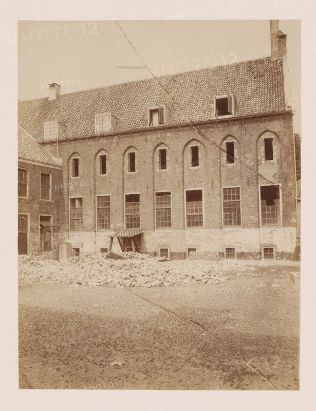 Former St. Agnieten Convent in Utrecht, photograph, annotation: 'Utrecht', annotation: 'Utrecht, Former St. Agnieten Convent now a military building, June 1902', Utrecht, 1902, photographer, Attributed to, paper, pasteboard, albumen print, height 219 mm x width 166 mm, cloisters, monastery, A sepia-toned photograph of the exterior wall of a multi-story brick building with a tiled sloping roof and several dormer windows, some open, and a chimney at one end; the facade shows arched window openings on the upper level and larger rectangular windows on the lower level, some with panes intact and some open or missing; in front of the building there is a pile of loose bricks and rubble, a small wooden lean-to structure against the wall, and a gravel-covered ground extending toward the foreground; a diagonal line or seam runs across the image from top to bottom.