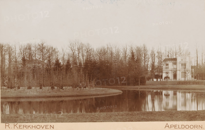 Oranjepark, Oranjepark in Apeldoorn, cabinet photograph, stamp: 'AF van Wieringen, Boek-, Muziek-, & Kunsthandel, Apeldoorn', collector's mark: 'Vosmaer', photographer: Richard Kerkhoven, Apeldoorn, vendor: AF van Wieringen, 1893 - 1902, Richard Kerkhoven, AF van Wieringen, photographer, vendor, paper, pasteboard, baryta paper, daylight collodion silver print, height 108 mm x width 168 mm, park, A calm body of water reflects a line of bare trees and a light-colored multi-story house with a tower-like section near the right edge of the scene; the house has prominent windows, a doorway, and an adjacent covered porch area, while the foreground shows a grassy bank and the water's edge, and the background is filled with tall leafless trees forming a continuous treeline across the image.