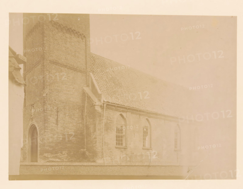 East side of the large church in Vollenhove, photograph, annotation: 'East side of the large church in Vollenhove (Overijsel) Aug 1898', annotation: 'Vollenhove', Vollenhove, 1898, photographer, Attributed to, paper, pasteboard, albumen print, height 171 mm x width 232 mm, church (exterior), A faded sepia-toned photograph of a brick building showing a tall tower corner and a sloped tiled roof, with arched windows set into the wall below the roofline and a narrow doorway at the tower base, a downspout running along the wall, and a short iron fence section visible near the windows.